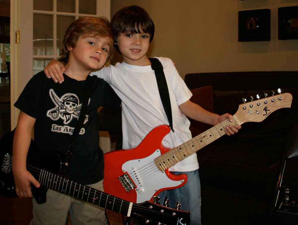 2 young boys stand holding guitars while one boy has his arm around the other boy's shoulders
